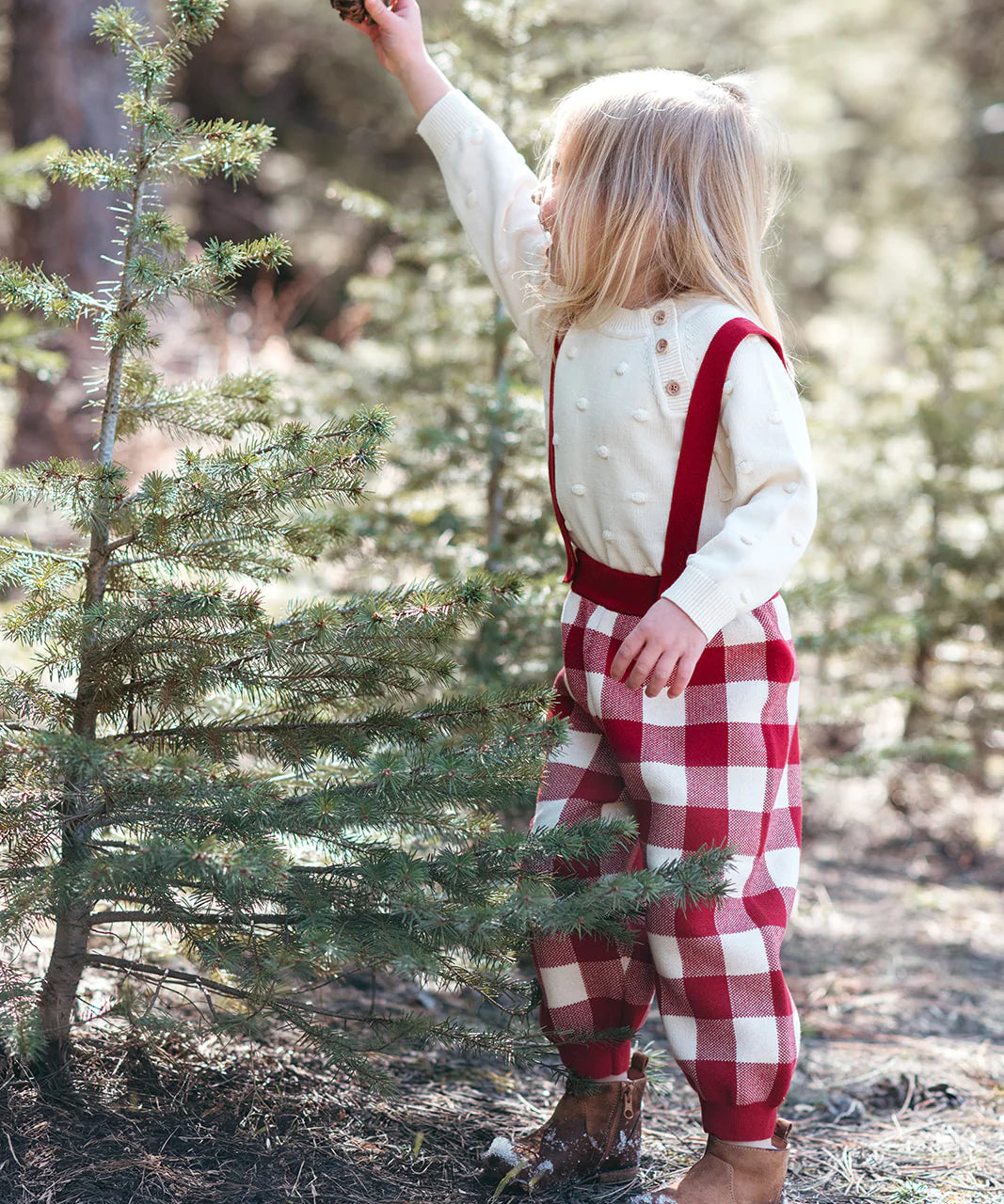 Child in a forest wearing red checkered overalls and a white sweater.
