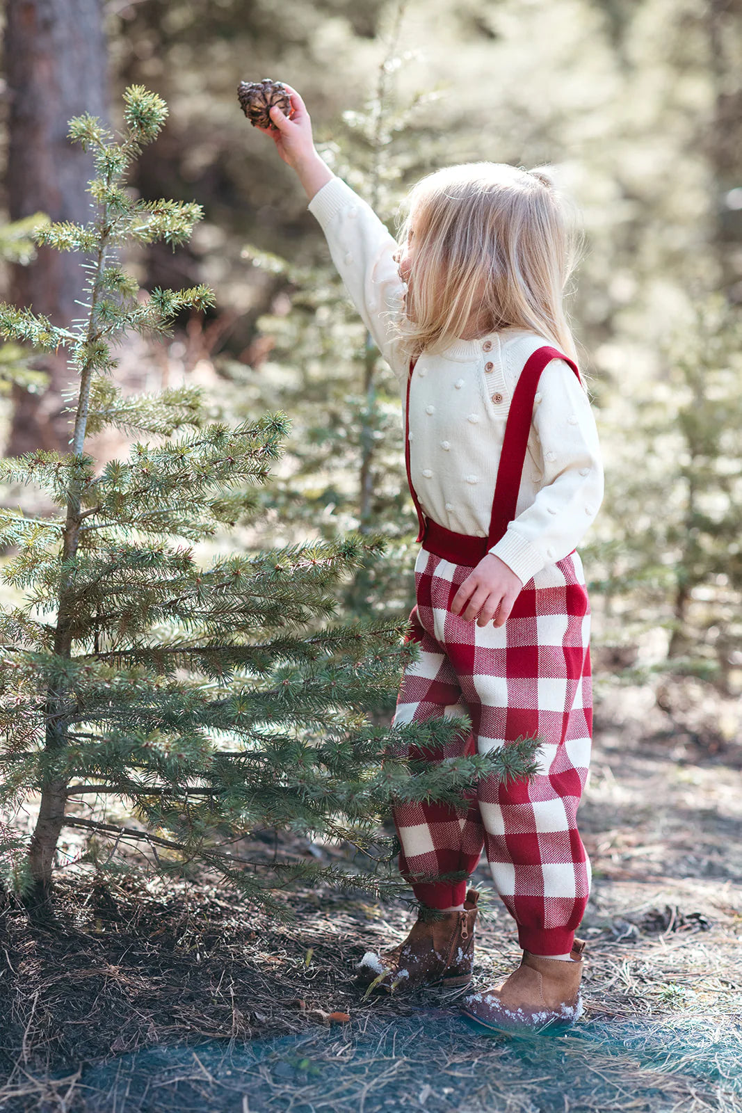 Child in a forest wearing red checkered overalls and a white sweater.